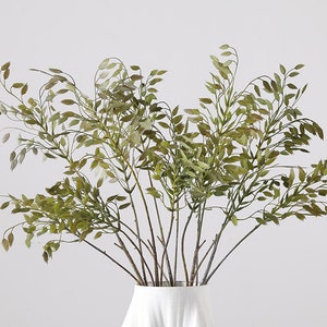 May include: A bouquet of dried green grass stems in a white vase. The stems are arranged in a loose, natural style, with the leaves cascading down the sides of the vase.