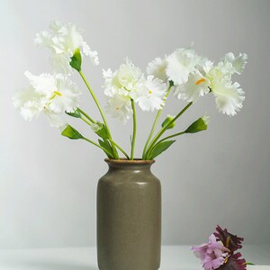 May include: A bouquet of white flowers with green stems in a brown ceramic vase. A single purple flower is on a white surface.