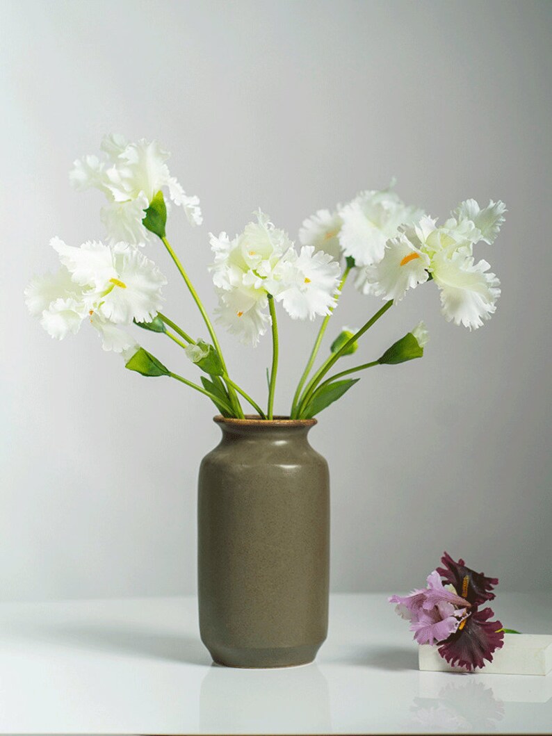 May include: A bouquet of white flowers with green stems in a brown ceramic vase. A single purple flower is on a white surface.