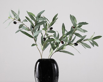 May include: A black vase with a green olive branch with black olives. The vase is round and has a smooth surface.