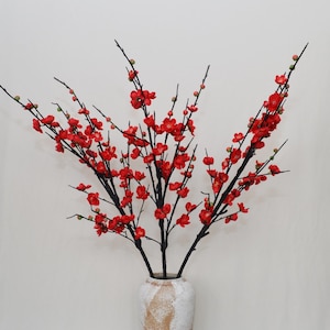 May include: A decorative floral arrangement with bright red flowers on dark stems. The stems are displayed in a cream-coloured vase. The arrangement is a decorative accent for home decor, adding a splash of colour.