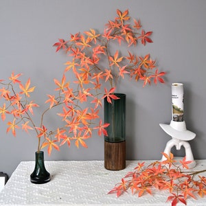 Puede incluir: Dos arreglos de ramas de arce artificiales con hojas naranjas y rojas en jarrones. El jarrón de la izquierda es de vidrio verde oscuro. El jarrón de la derecha es un cilindro verde oscuro con una base de madera marrón.