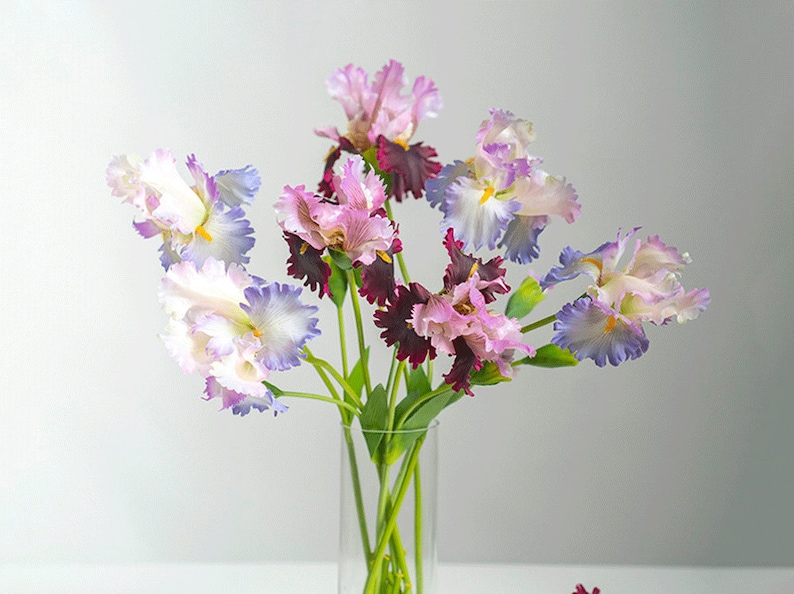 May include: A bouquet of purple, pink, and white irises in a clear glass vase. The irises have ruffled petals and are arranged in a vase on a white surface.