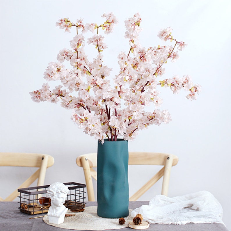 May include: A bouquet of delicate pink cherry blossom branches arranged in a teal blue textured vase. The vase is on a table with a white tablecloth and a wooden chair in the background.