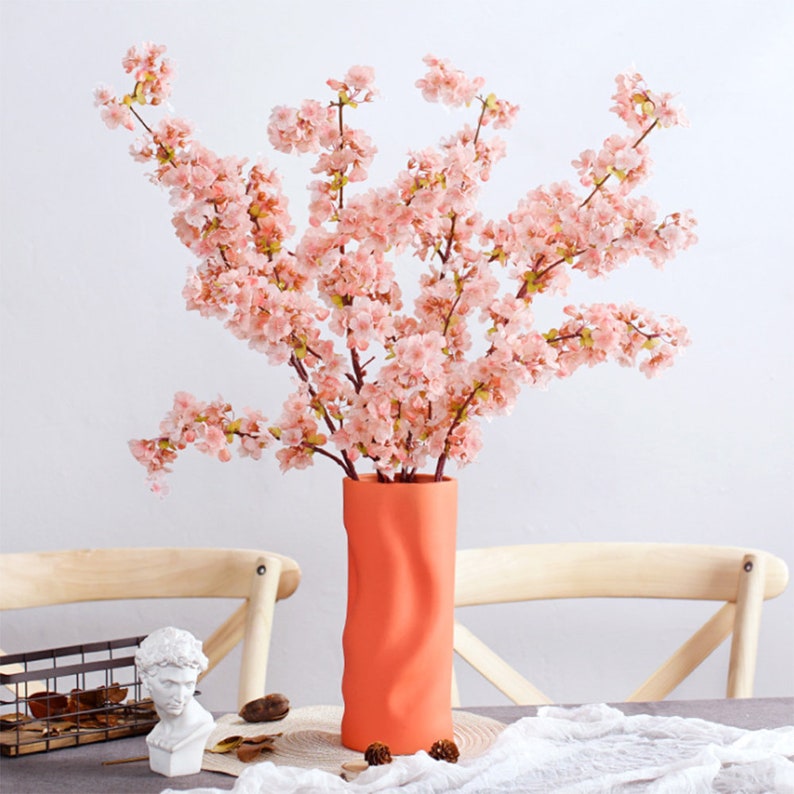 May include: A bouquet of pink cherry blossoms in a bright orange, textured ceramic vase. The vase has a wavy, rippled design.