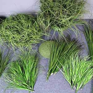 May include: An assortment of artificial green grass and plants. The image features several bundles of long, slender, bright green grass, along with a round, moss-covered sphere and a woven basket with the word "Plants" on it.