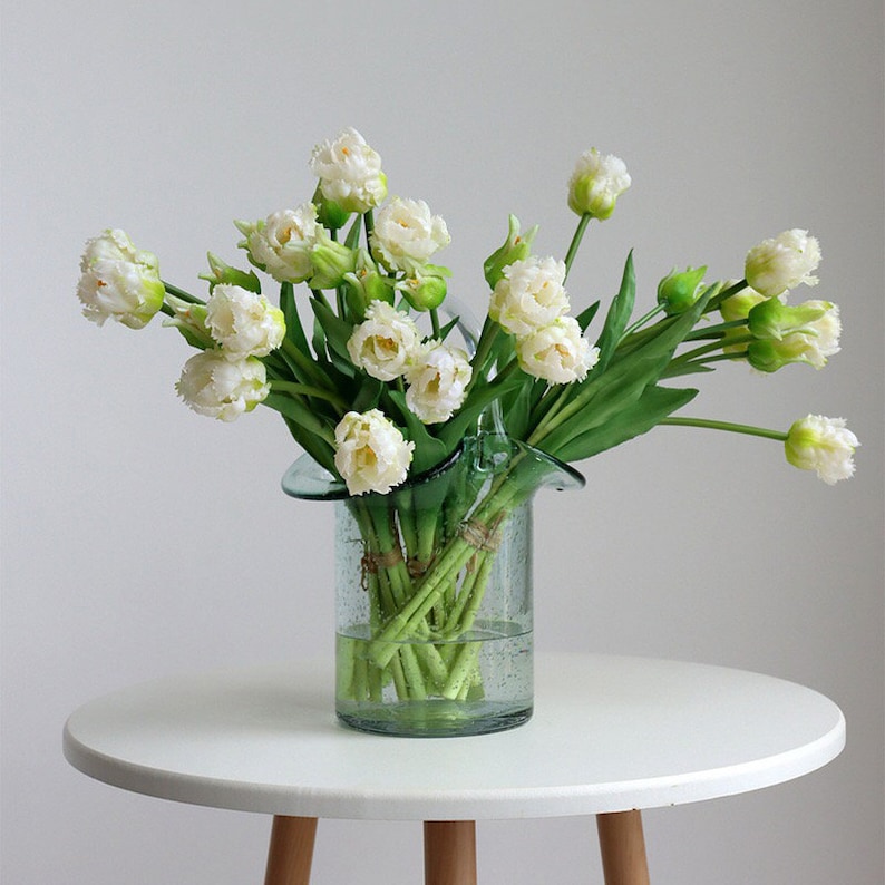 Op de afbeelding: Een boeket witte tulpen gerangschikt in een heldere glazen vaas met een golvende rand. De vaas staat op een witte tafel met houten poten.