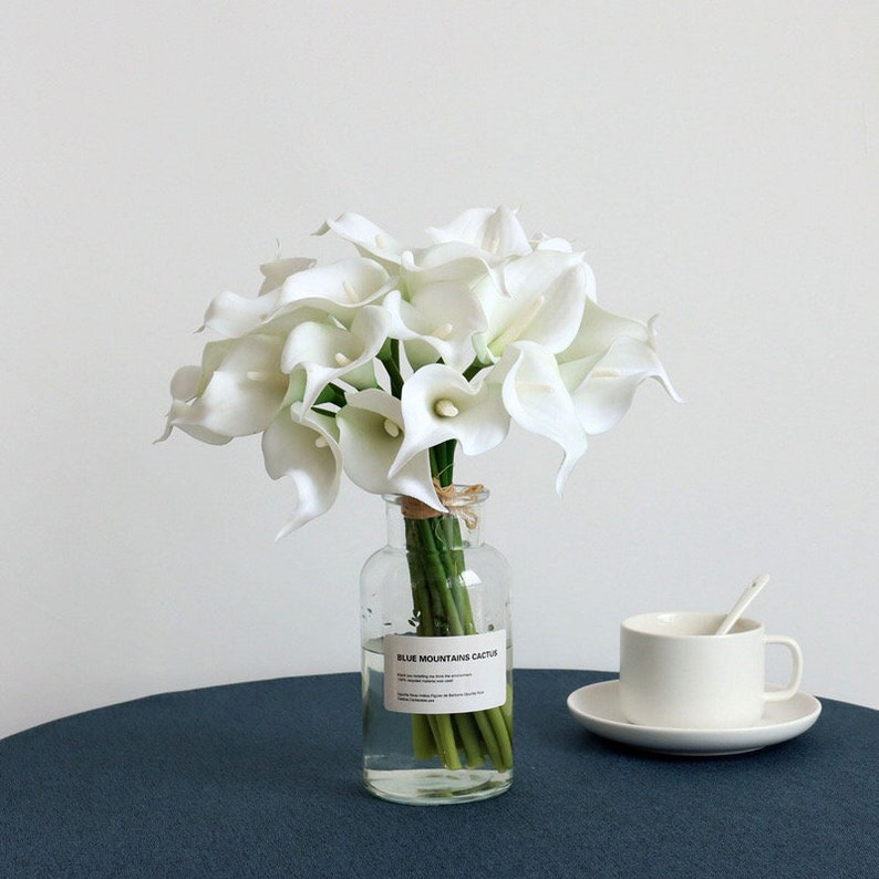 May include: A bouquet of white calla lilies in a clear glass vase with a label that reads "Blue Mountains Cactus".