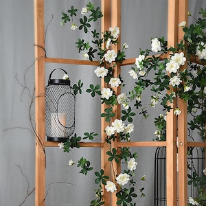 May include: A black metal lantern with a wire mesh design hangs from a wooden trellis with white flowers and green leaves growing around it.