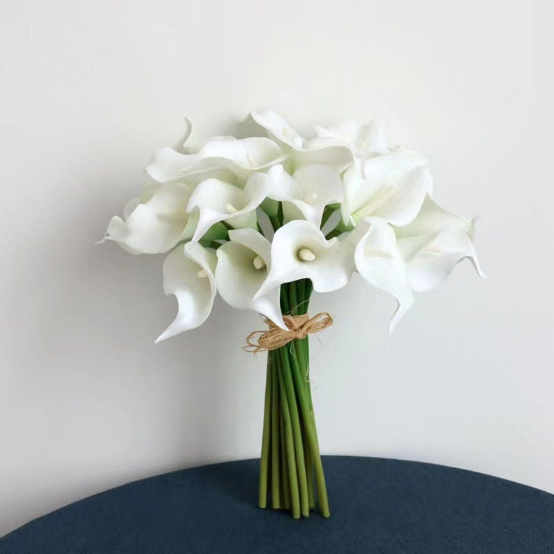May include: A bouquet of white artificial calla lilies tied together with twine. The flowers are arranged in a cluster and have a realistic appearance.