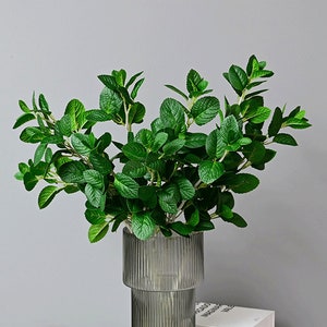 May include: A bunch of artificial green leafy plants in a clear glass vase with a textured surface. The vase is sitting on a white surface.