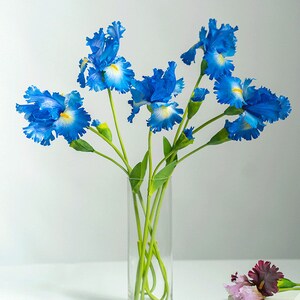 May include: A bouquet of blue irises with yellow centers in a clear glass vase. The flowers are arranged in a vase on a white surface.