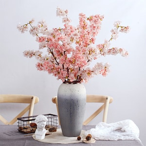 May include: A bouquet of pink and white artificial cherry blossoms in a gray ceramic vase with a white and gray ombre effect. The vase is sitting on a table with a wooden chair and a white cloth.