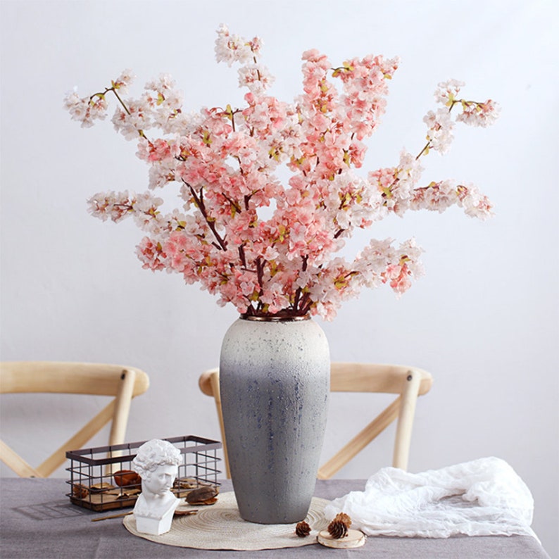 May include: A bouquet of pink and white artificial cherry blossoms in a gray ceramic vase with a white and gray ombre effect. The vase is sitting on a table with a wooden chair and a white cloth.