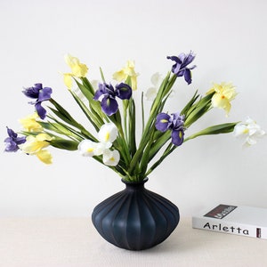 May include: A bouquet of purple, white, and yellow irises in a dark blue vase. The vase has a ribbed design. The flowers are arranged in a vase on a white surface.