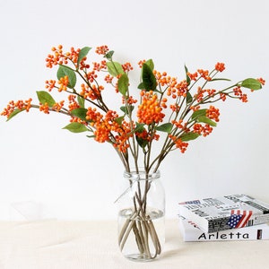 May include: A bouquet of orange berry branches with green leaves in a clear glass vase. The vase is sitting on a table with a book titled "Arletta" on top of it.