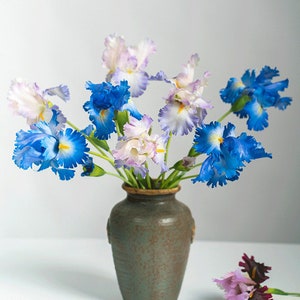 May include: A bouquet of blue, white, and purple irises in a green ceramic vase. The vase has a textured surface and is sitting on a white surface.