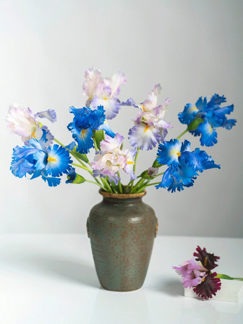 May include: A bouquet of blue, white, and purple irises in a green ceramic vase. The vase has a textured surface and is sitting on a white surface.