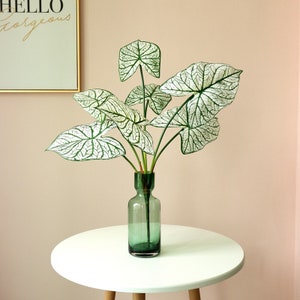 May include: A green glass vase holding artificial Caladium leaves with white and green veining. The vase sits on a white table with wooden legs. A framed print with the words "HELLO gorgeous" is in the background.