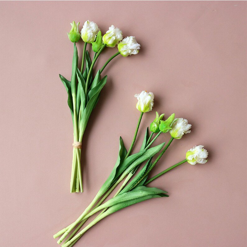 Op de afbeelding: Twee bosjes witte kunsttulpen met groene stelen en bladeren. De tulpen zijn gerangschikt op een lichtbruine achtergrond.