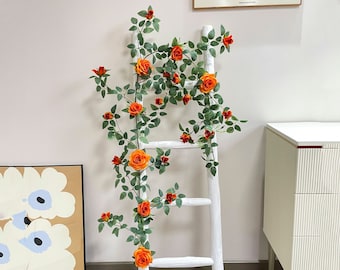 Puede incluir: Una escalera blanca con una guirnalda de rosas artificiales de color naranja y verde envuelta alrededor. La guirnalda está hecha de plástico y tiene un aspecto realista.