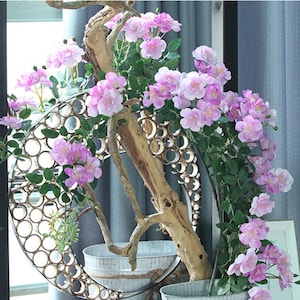 Puede incluir: Arreglo floral decorativo con flores de color rosa claro y hojas verdes, dispuestas alrededor de una rama de madera desgastada. El arreglo se encuentra dentro de un marco circular de metal con una serie de agujeros y en un cubo de metal.