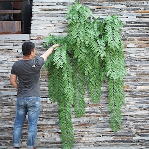 May include: A green artificial hanging plant with long, leafy vines against a gray stone wall.