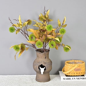 May include: A brown ceramic vase with a white deer cutout, filled with a bouquet of artificial fall foliage with green and brown burrs.