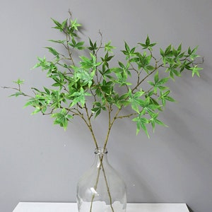 May include: A green artificial maple branch with multiple stems and leaves in a clear glass vase.