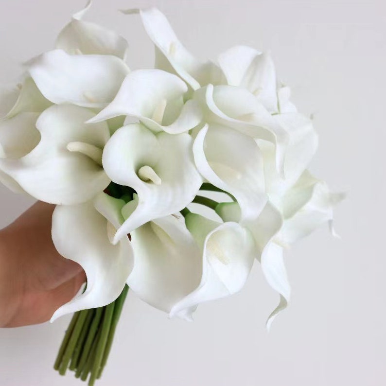 May include: A bouquet of white calla lilies with green stems. The flowers are arranged in a cluster and are ready to be used for a wedding or other special occasion.