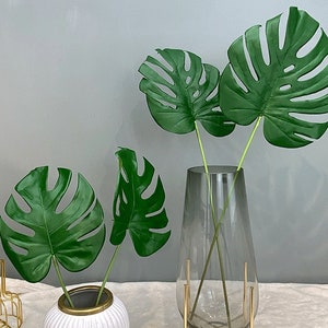 May include: Three artificial monstera leaves in a clear glass vase with a gold metal stand. The vase is on a white surface.