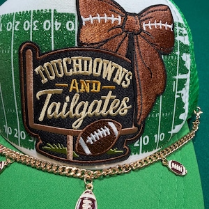 May include: A green and white trucker hat featuring a glittery football field pattern and a large embroidered patch that reads Touchdowns and Tailgates. The hat is decorated with a brown bow and a gold chain with small football charms attached.