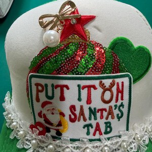 May include: A white and green trucker hat featuring a rectangular patch with the text "PUT IT ON SANTA'S TAB" and a Santa Claus graphic. The hat is decorated with a red sequined tree, a gold bow brooch with a pearl, and a white lace trim with pearls.