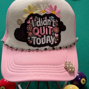 May include: A light pink and white trucker hat featuring a black patch with the embroidered text I didn't QUIT TODAY surrounded by colorful flowers and hearts. The hat is accented with a silver-toned rhinestone chain and a floral pearl embellishment.