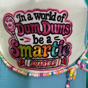 May include: A light blue trucker hat with a white front panel featuring an embroidered patch that reads In a world of Dum Dums be a Smartie with a graphic of a lollipop and a candy roll. The hat is decorated with a string of colorful beads.