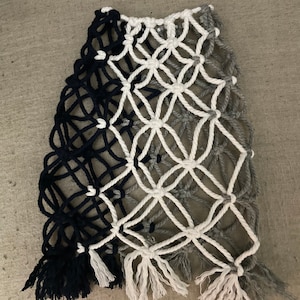 以下が含まれることがあります： フリンジが付いた黒、白、グレーの壁掛けマクラメ。マクラメは太いロープで作られており、幾何学模様になっています。