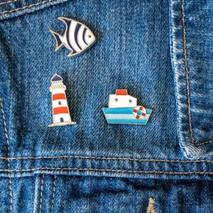 Set of 3 nautical magnetic enamel pins - lighthouse, fish and boat - worn on a denim jacket.