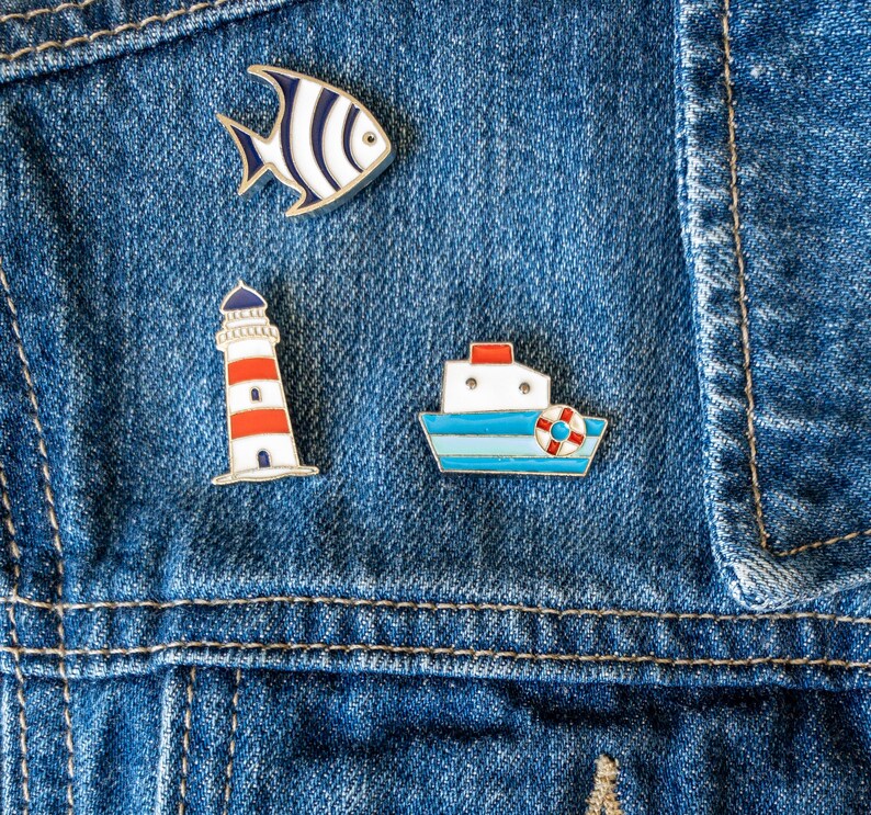 Set of 3 nautical magnetic enamel pins - lighthouse, fish and boat - worn on a denim jacket.