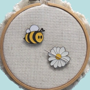 May include: A white daisy and a yellow and black bee pin on a beige fabric background. The pins are displayed in a wooden embroidery hoop.