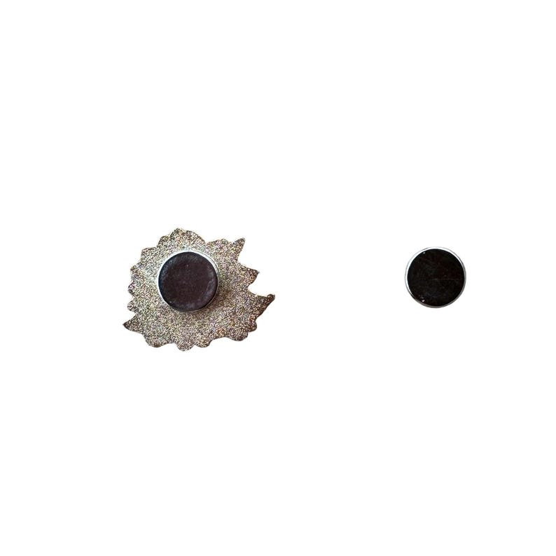 May include: A silver-colored magnet in the shape of a leaf with a dark brown circular magnet attached. A separate dark brown circular magnet is also visible. The items are on a white background.