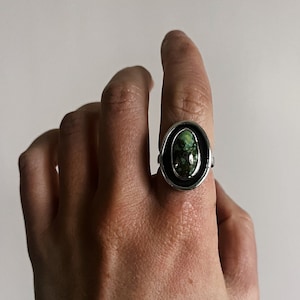 May include: A silver ring with a green oval gemstone set in a black bezel. The ring is on a person's finger.