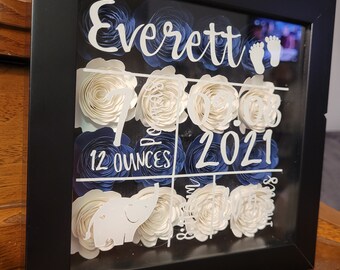 May include: A black shadow box with white paper flowers and a white elephant cutout. The text "Everett" and "7.09.2021" is visible in the box. The text "12 ounces" is also visible.