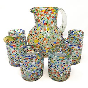 May include: A colorful glass pitcher with a handle and six matching glasses. The glassware is decorated with a speckled pattern of red, blue, yellow, and green dots. One glass contains a lime wedge.