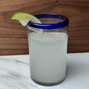 May include: A tall glass with a blue rim filled with a white liquid and a lime wedge garnish. The glass is sitting on a white marble surface.