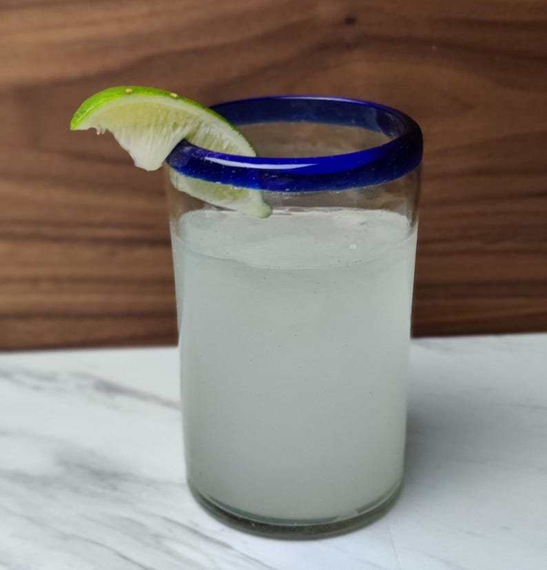 May include: A tall glass with a blue rim filled with a white liquid and a lime wedge garnish. The glass is sitting on a white marble surface.