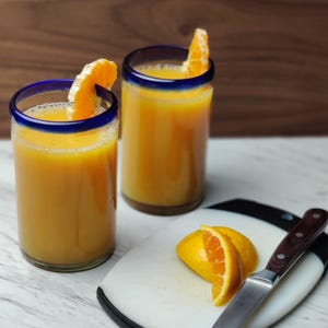 May include: Two glasses of orange juice with orange slices on the rim. The glasses have a blue rim and are sitting on a white marble surface. There is a white cutting board with a knife and orange slices on it.