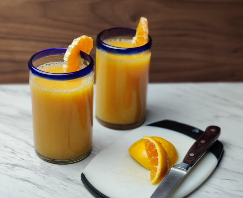 May include: Two glasses of orange juice with orange slices on the rim. The glasses have a blue rim and are sitting on a white marble surface. There is a white cutting board with a knife and orange slices on it.