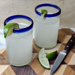 May include: Two clear glass tumblers with blue rims filled with a white liquid and garnished with lime wedges. The glasses are sitting on a wooden cutting board with a brown handled knife.