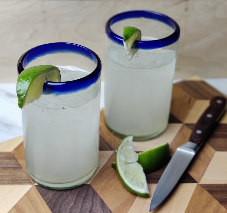 May include: Two clear glass tumblers with blue rims filled with a white liquid and garnished with lime wedges. The glasses are sitting on a wooden cutting board with a brown handled knife.