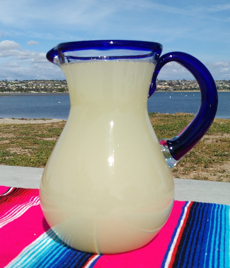 May include: A clear glass pitcher with a blue rim and handle, filled with a light yellow liquid. The pitcher is sitting on a colorful striped blanket.